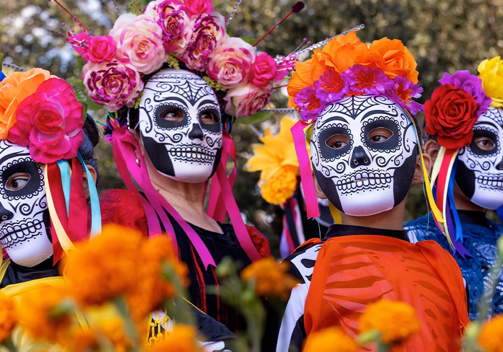 People wearing Day of the Dead masks and floral headpieces stand together, surrounded by vibrant marigolds during a festival in an outdoor setting with trees in the background.