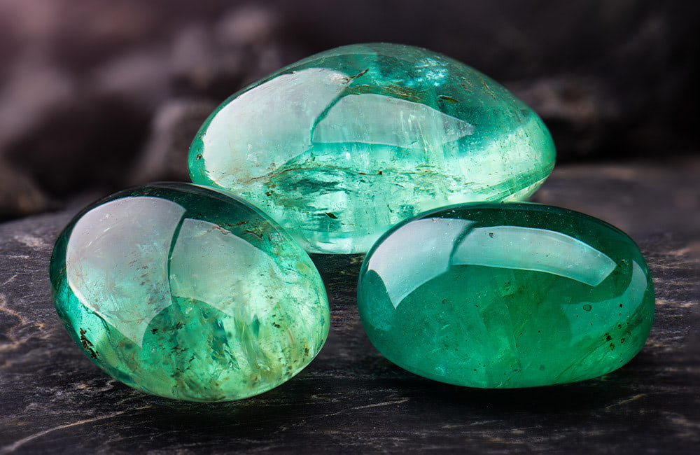 Three polished green gemstones resting on a dark, textured surface. The stones are smooth and semi-transparent, reflecting light in a dimly lit setting.