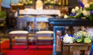 Flower arrangement with white and blue flowers adorns a wooden pew. In the background, two benches face a decorated altar covered with a white cloth, in a church setting.