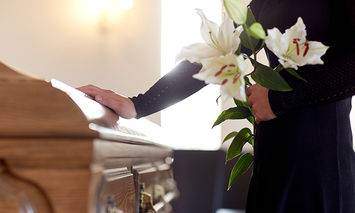 A person dressed in black holds white lilies beside a wooden coffin with polished handles, inside a softly lit room.