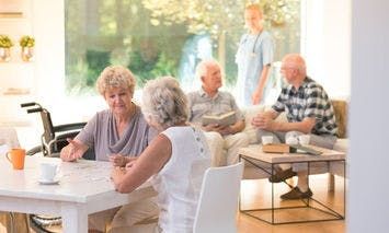 Elderly women sit at a table in conversation; two men in armchairs chat; a caregiver stands in a well-lit, home-like common area.