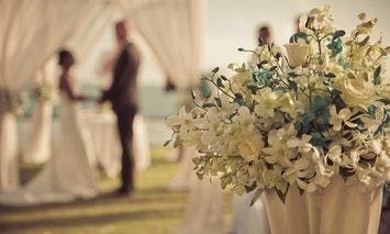 A bouquet of white and green flowers sits in the foreground, while a bride and groom exchange vows under a decorated canopy in a sunny outdoor setting in the blurred background.